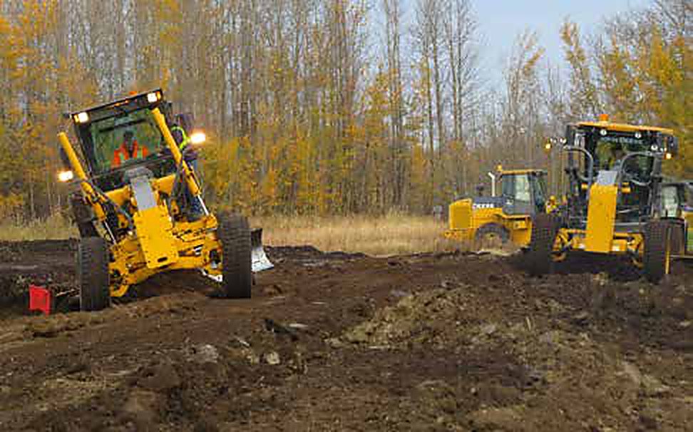 Motor graders working a gravel road corridor cut through standing forest.